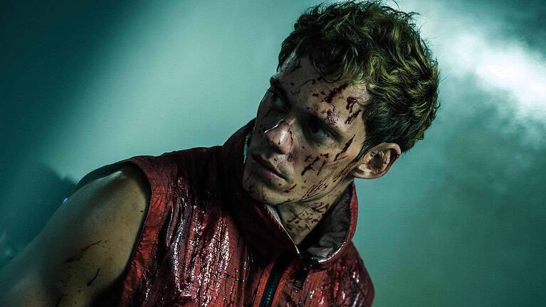 A young man with bloody and dirt-stained skin appears exhausted, his expression intense and alert in a dimly lit, foggy background.