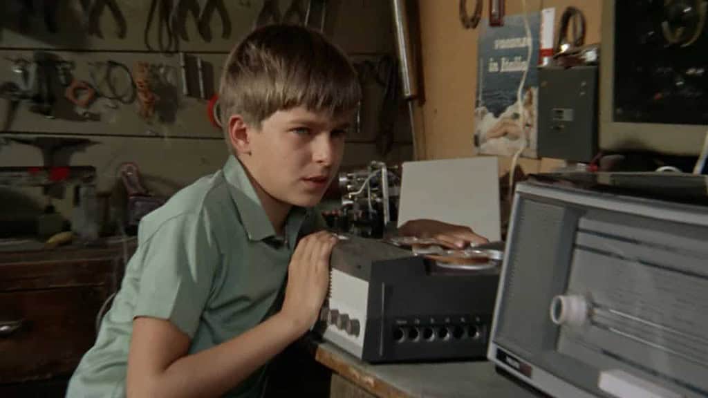 A young boy in a workshop listening intently to a reel-to-reel tape recorder, surrounded by various tools hanging on the wall.
