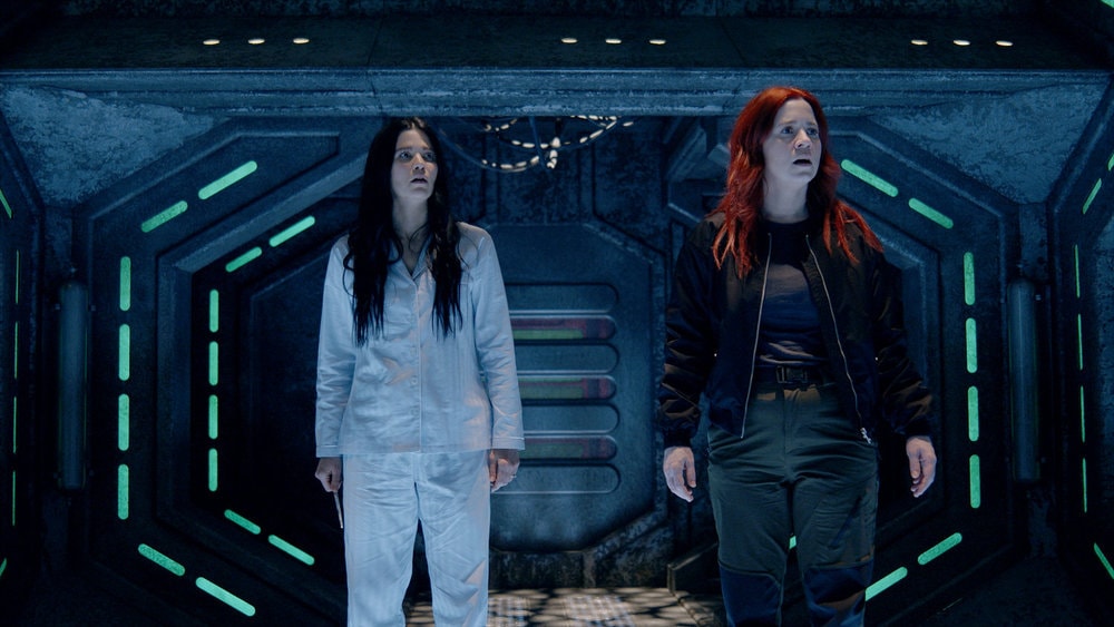Two women with apprehensive expressions standing inside a dimly lit futuristic corridor with blue lighting accents.