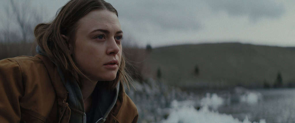 A woman with a contemplative expression looks into the distance, wearing a brown jacket, against a blurred natural landscape with water and hills.