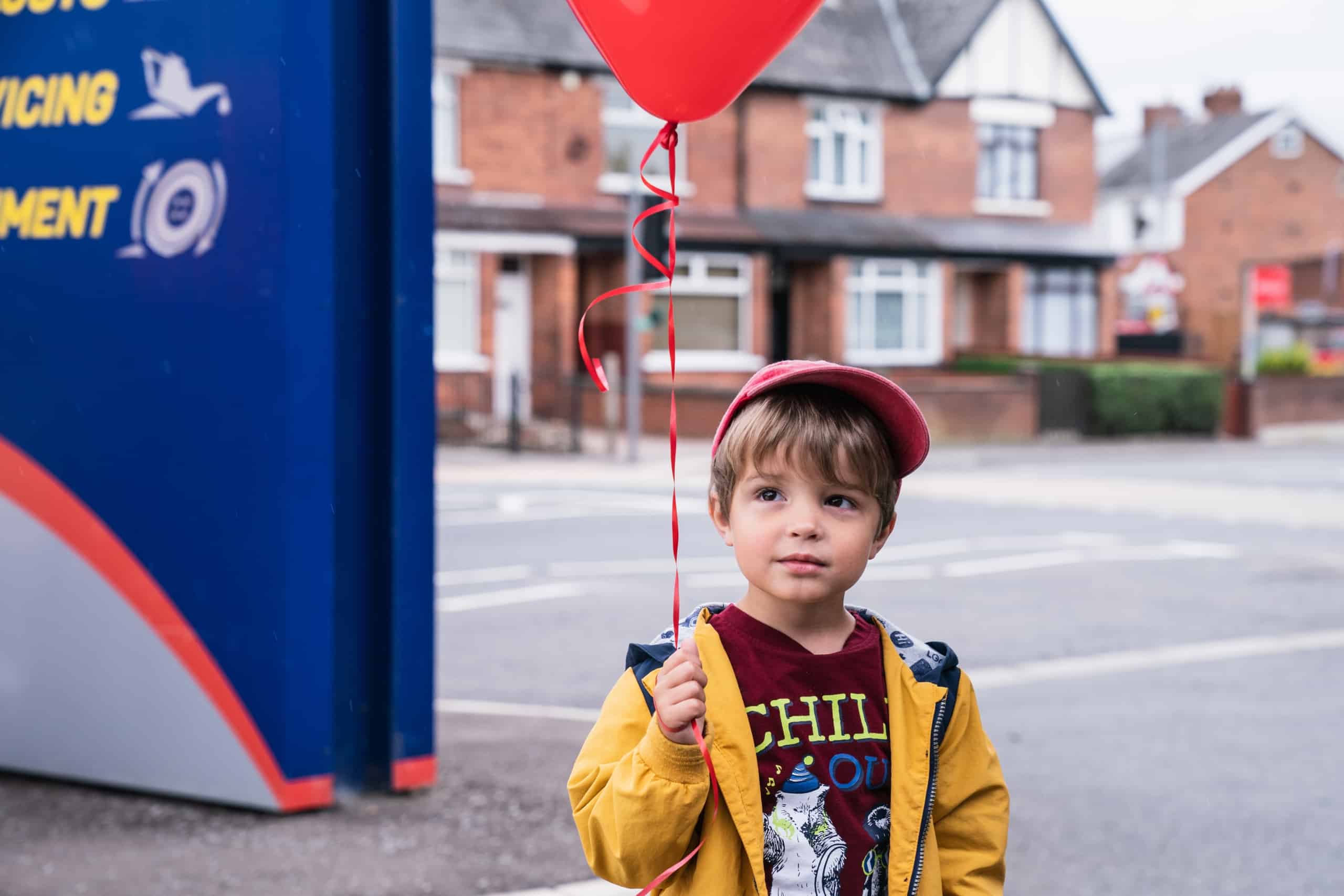 A young child wearing a red hat and yellow backpack holds a red balloon, standing on a sidewalk by a blue sign.