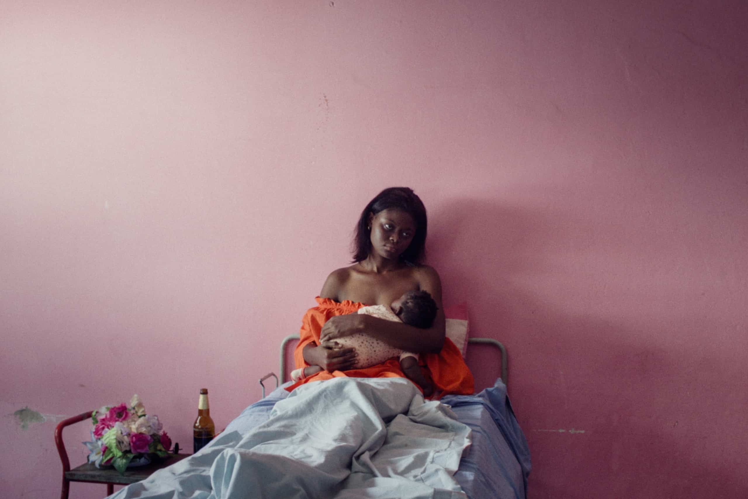 A woman sitting on a bed, feeding her baby and looking directly at the camera, in a room with pink walls and minimal furnishings.