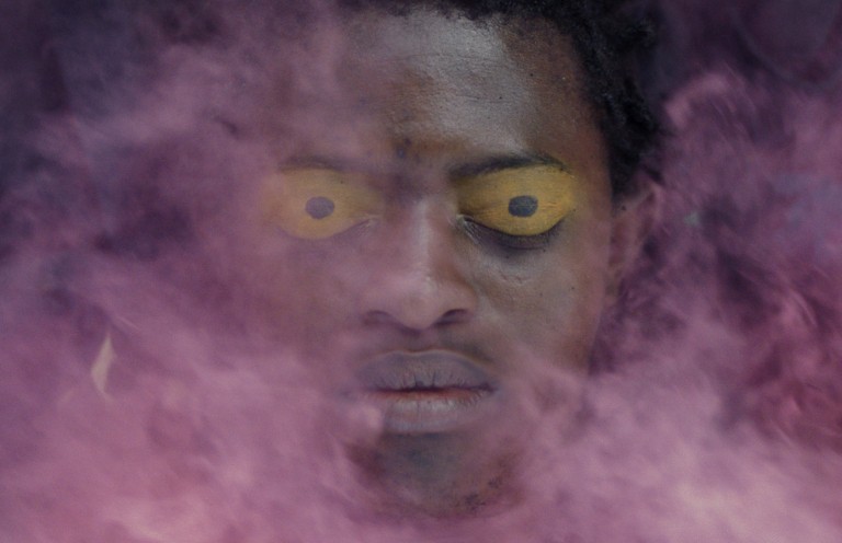 A man with striking yellow eyes appears through a dense purple smoke, creating a mysterious aura.