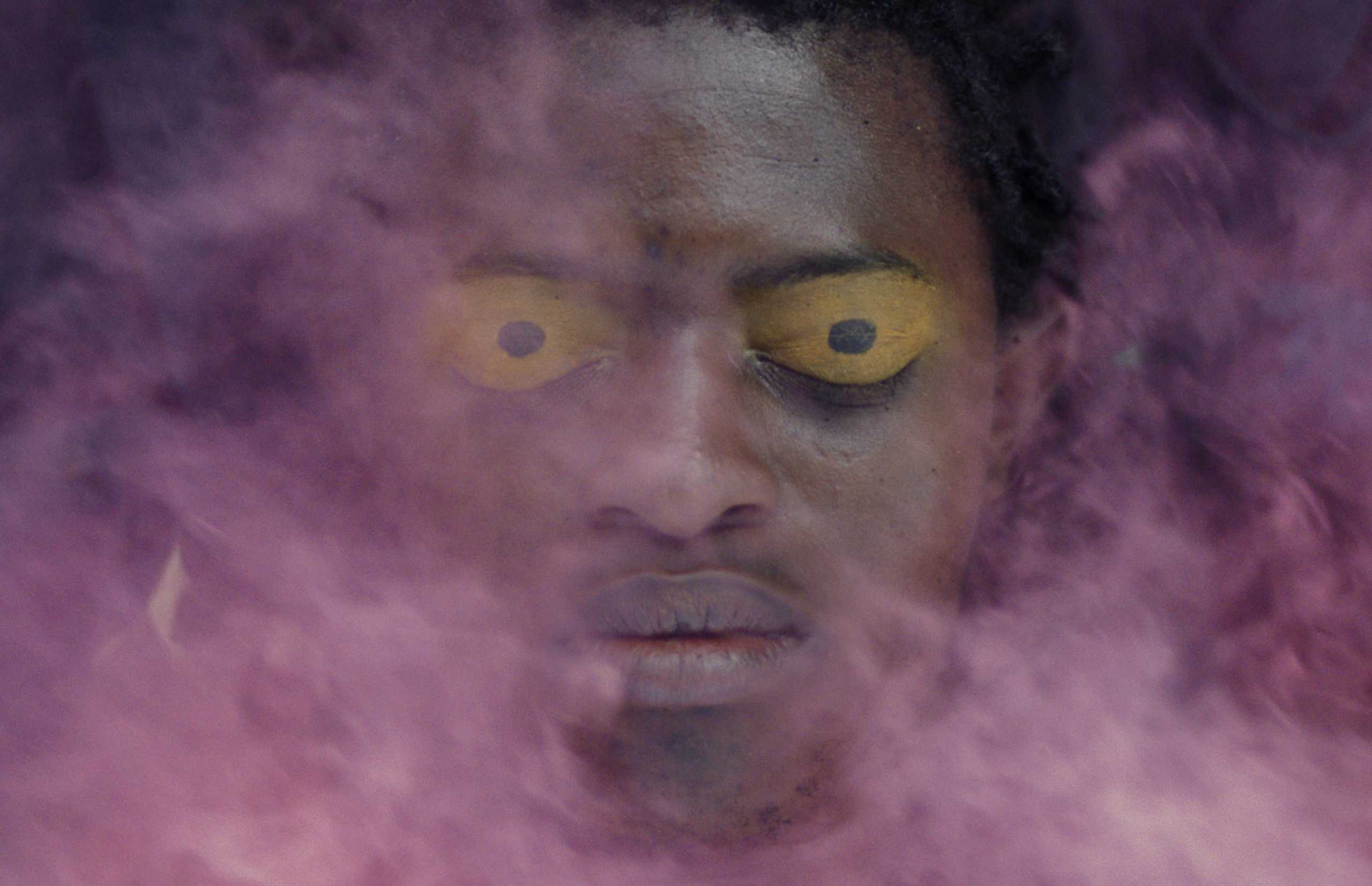 A man with striking yellow eyes appears through a dense purple smoke, creating a mysterious aura.