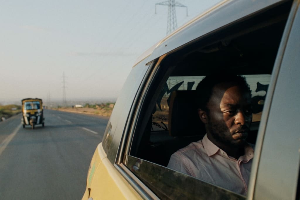 A focused man driving a car on a highway, seen through the window with a sunset-lit rural landscape in the background.