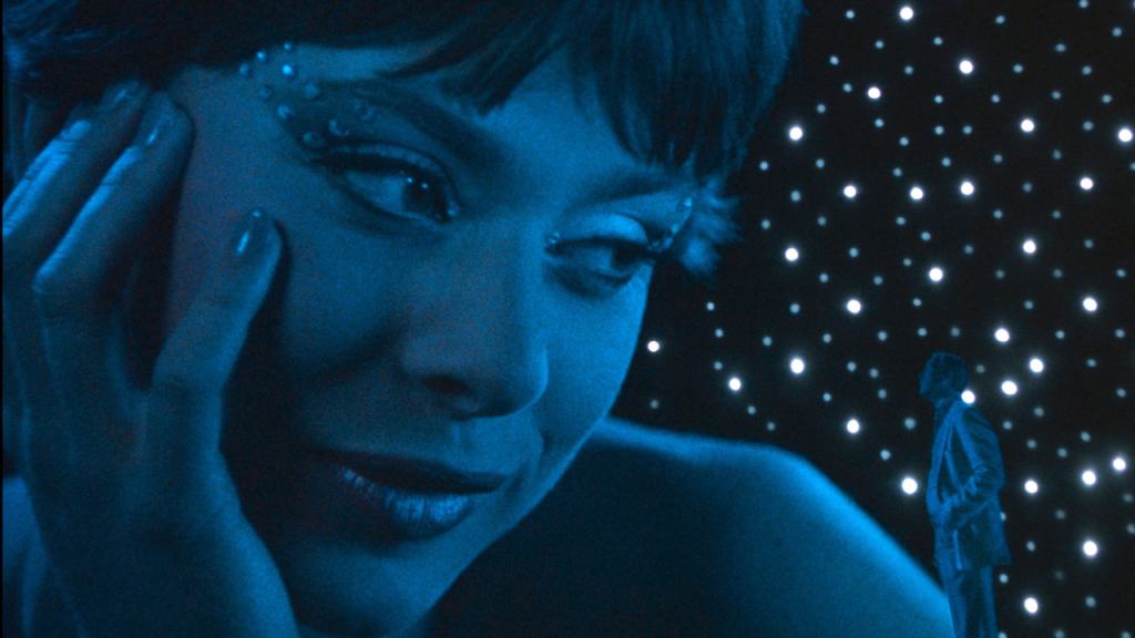 A close-up of a smiling person with a face adorned with gemstones, against a backdrop of a starry scene with a small silhouette of another figure.