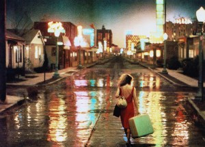 A woman in a red dress walking down a wet street at night, lined with neon signs.