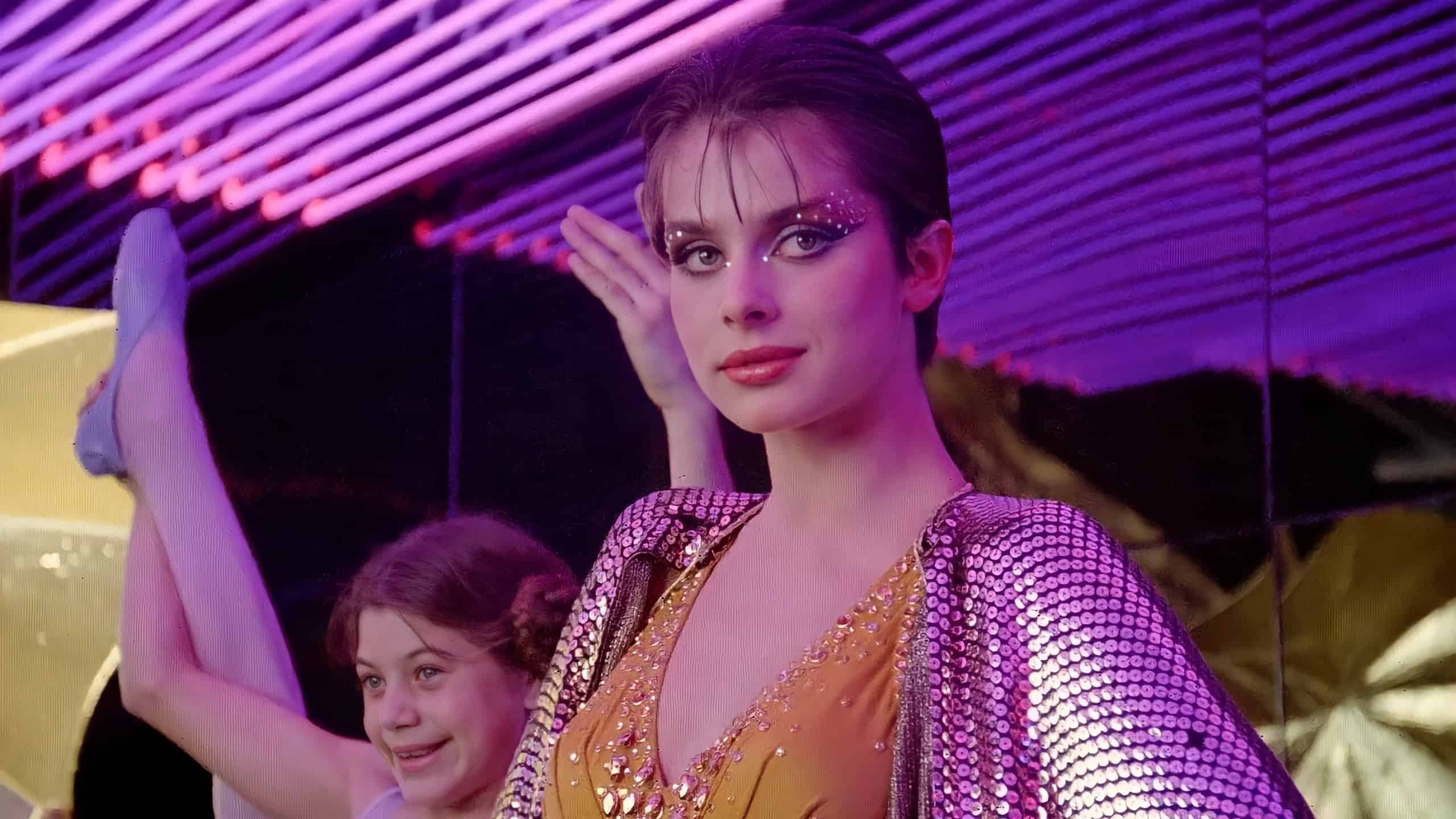 A woman in a sequined costume with makeup and rhinestones poses confidently, accompained by a smiling girl in the background under pink neon lights.