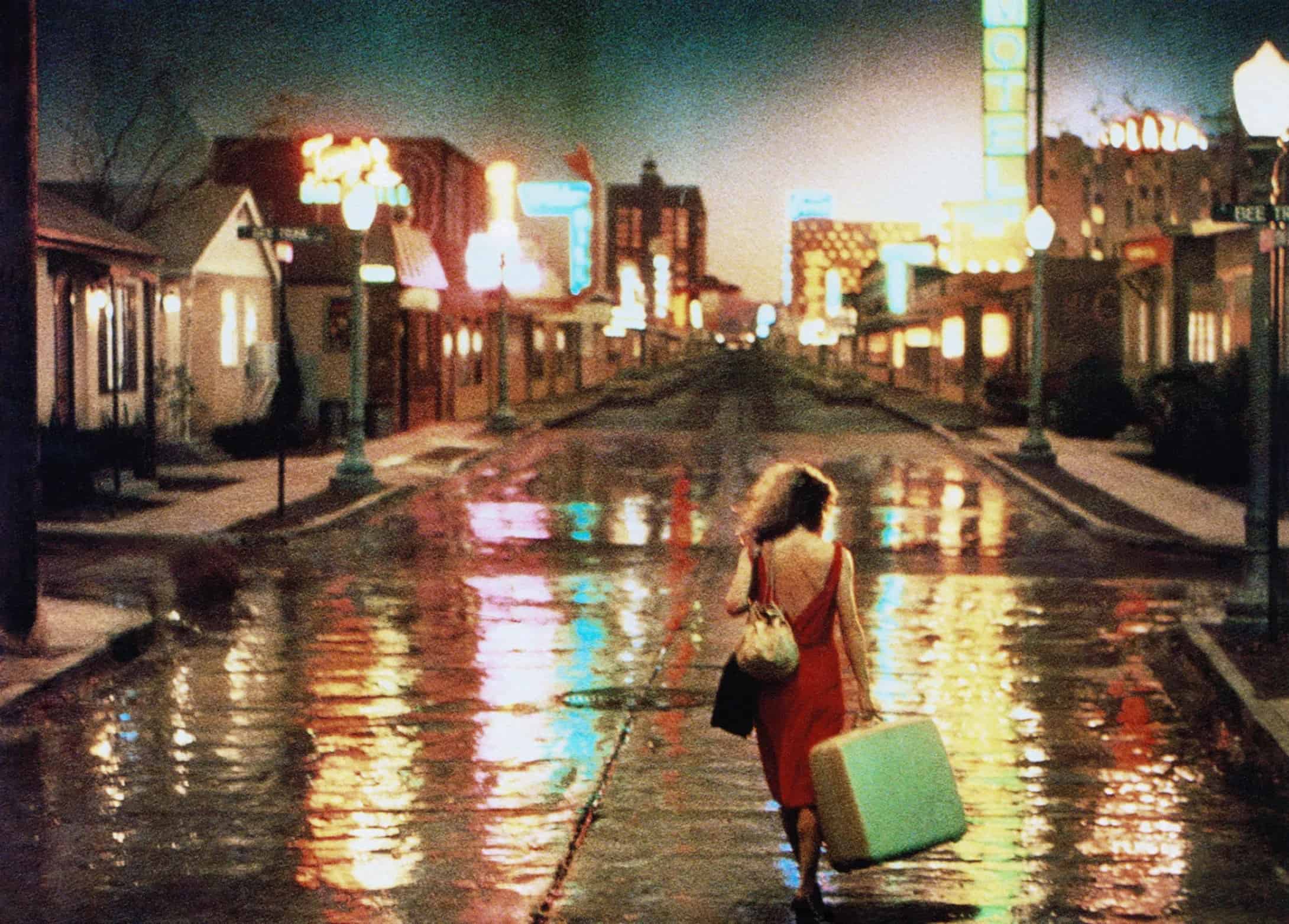 A woman in a red dress walking down a wet street at night, lined with neon signs.