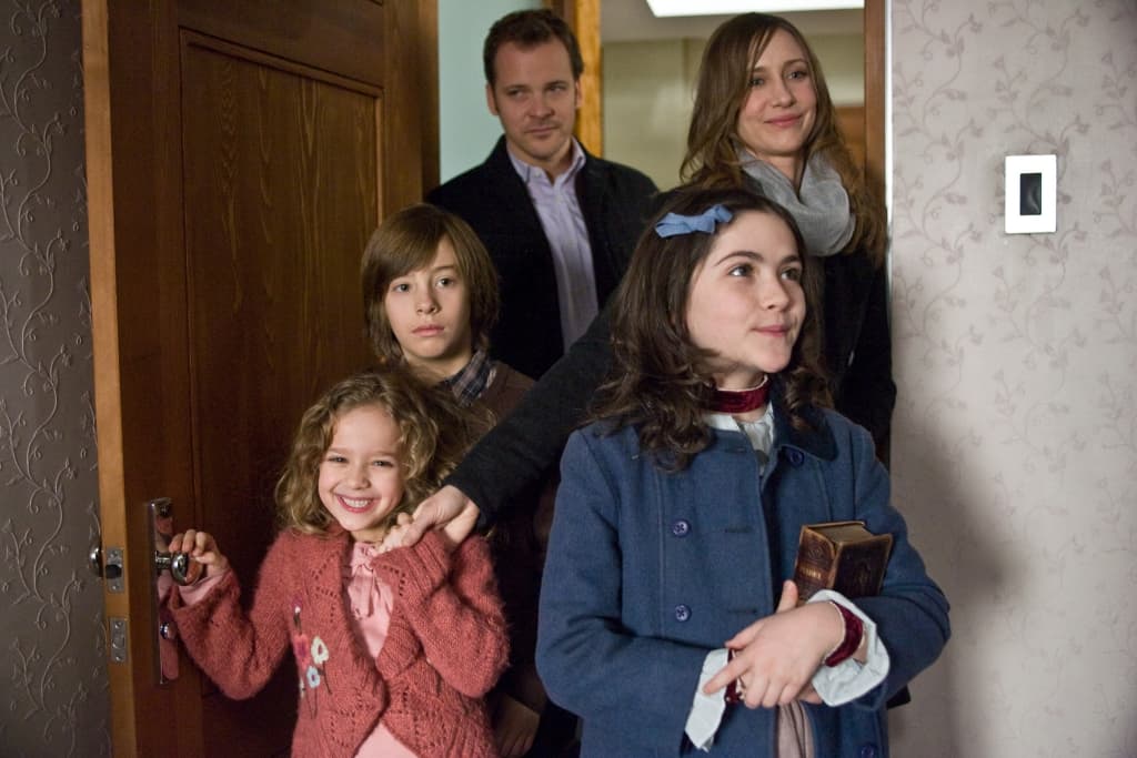 A family with two adults and three children standing at a doorway, looking towards something outside of the frame with expressions of anticipation or curiosity.