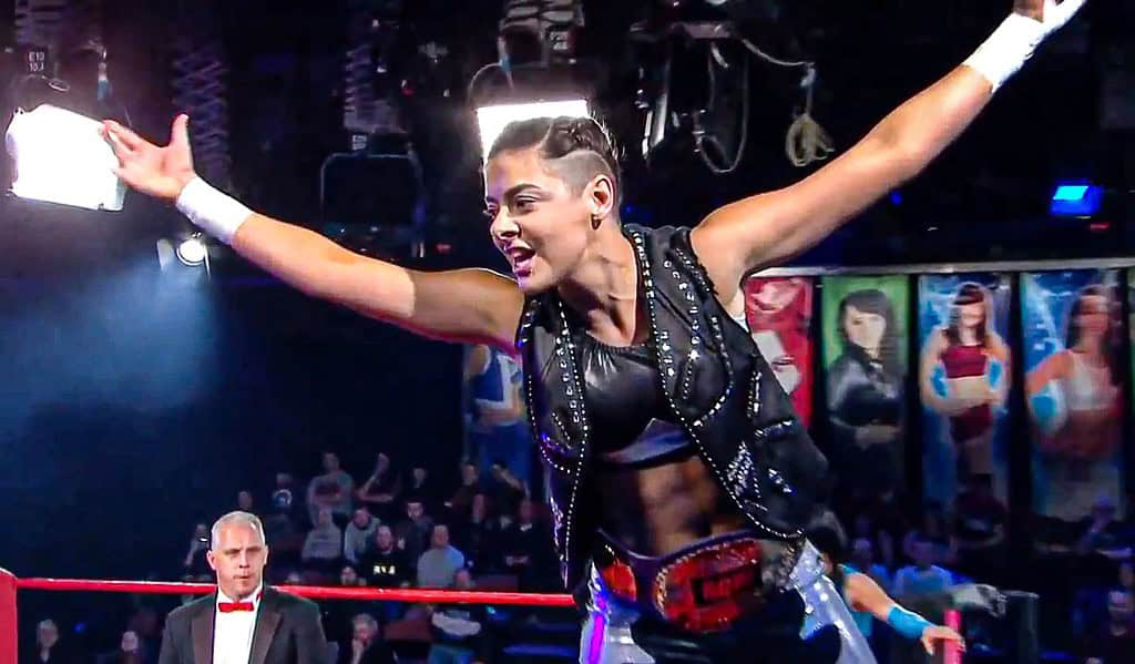 A professional wrestler celebrating in the ring with a championship belt, arms raised, in front of a cheering crowd and a focused referee in the background.