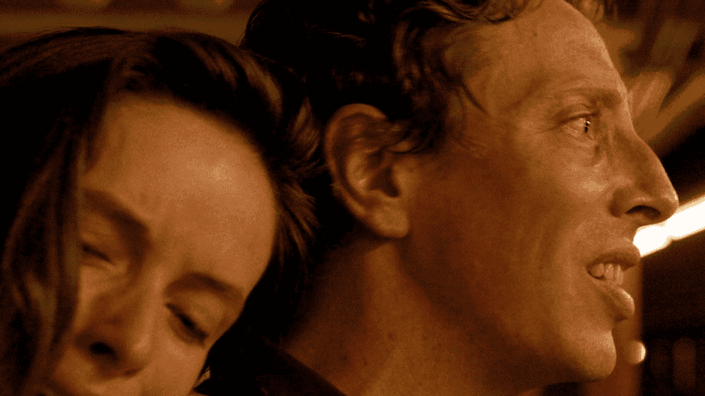 A close-up of a man and woman looking to the right with thoughtful expressions, illuminated in low light.