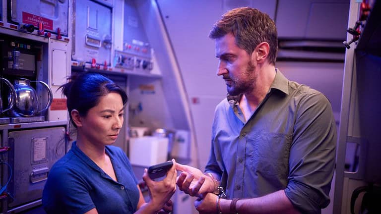 ‘Red Eye’ Series 1 Review – Not The Thrilling Highflyer It Wants To Be Two people, a woman and a man, examine a smartphone together in a dimly lit room filled with technical equipment.