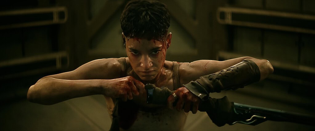 A bloodied man intensely grips a bow, aiming an arrow, in a dimly lit industrial setting.