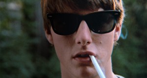 Close-up of a young man wearing sunglasses and a cigarette in his mouth, with a blurred green background.