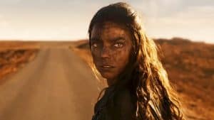 A woman with a dirt-covered face stands on a desert road, looking intently forward.