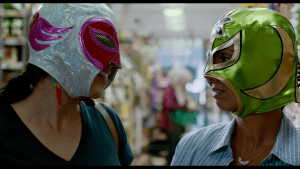 Two people wearing colorful luchador masks facing each other indoors.
