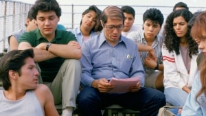 A group of students listening attentively to a man reading from a paper outdoors.