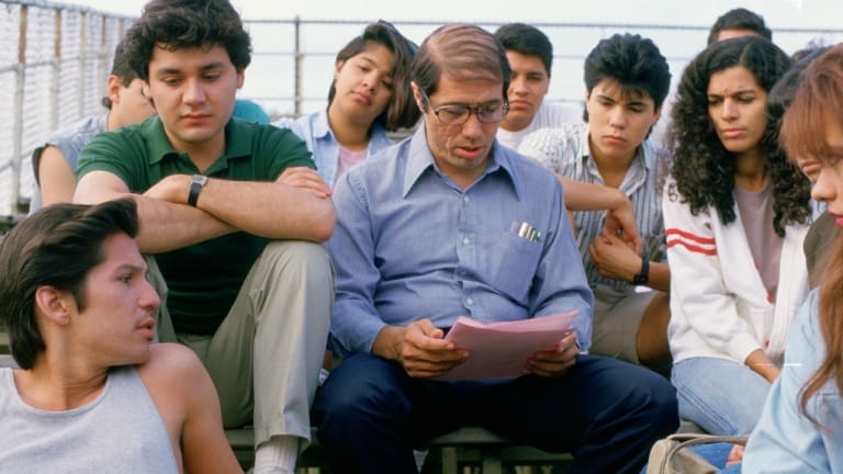 A group of students listening attentively to a man reading from a paper outdoors.