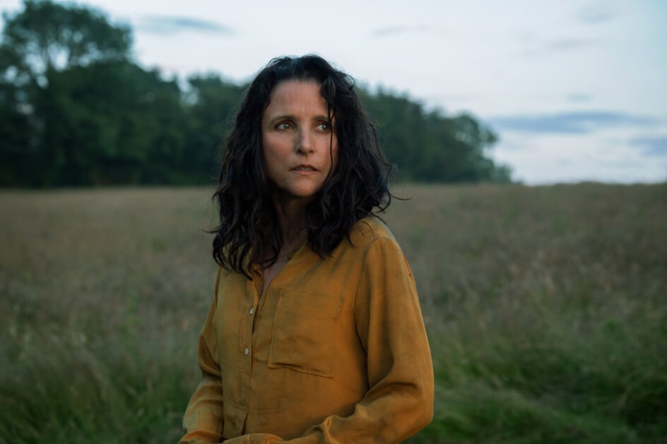 Woman with long dark hair wearing a mustard-colored shirt stands in a grassy field with trees in the background.