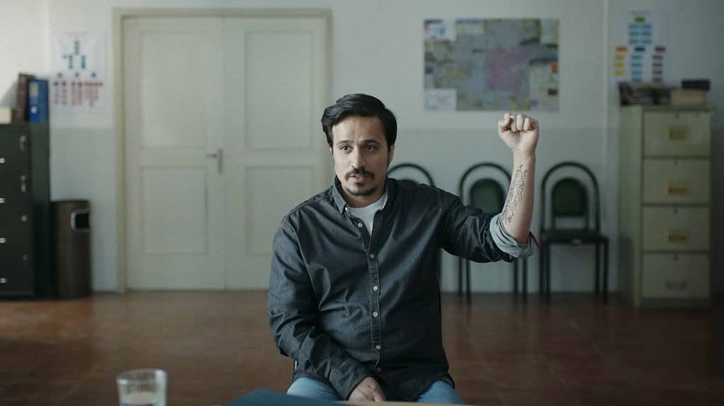 A man seated in an office, raising his fist, with a clock and maps on the wall behind him.