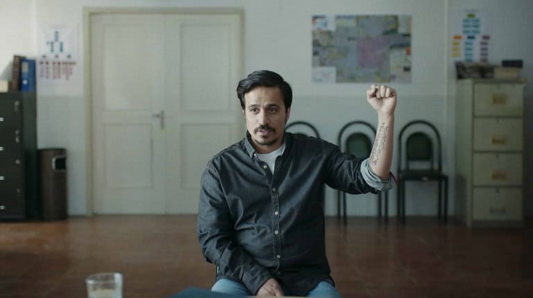A man seated in an office, raising his fist, with a clock and maps on the wall behind him.