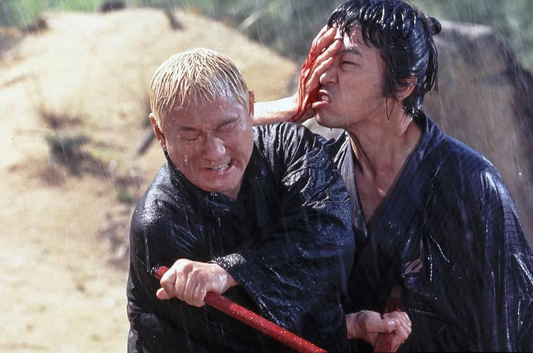 Two men engaged in a martial arts fight in the rain, with one man wielding a red staff and striking the other in the face.