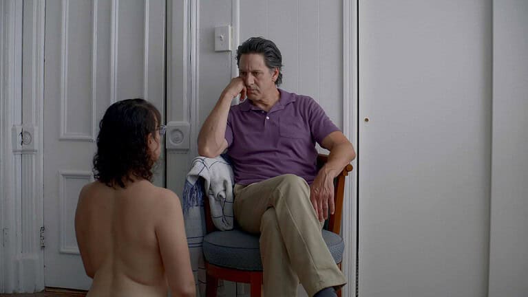 ‘The Feeling That The Time For Doing Something Has Passed’ Review – Irony Or Apathy? A man in a purple shirt seated on a chair listens intently to a child, who is seated facing him, only the back of the child visible. they are in a room with a white door and wall.