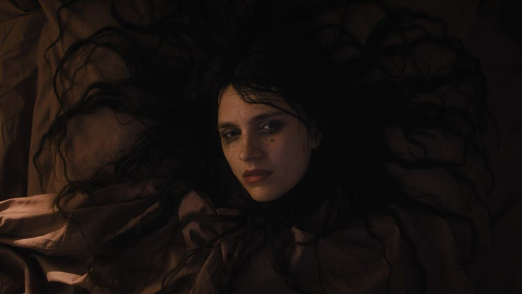 A woman lying down with her dark hair spread out, looking at the camera with a somber expression.