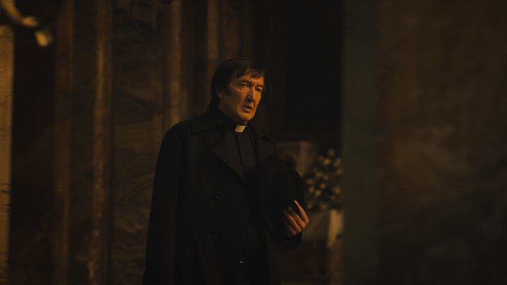 A man dressed as a priest stands in a dimly lit room, holding a hat.