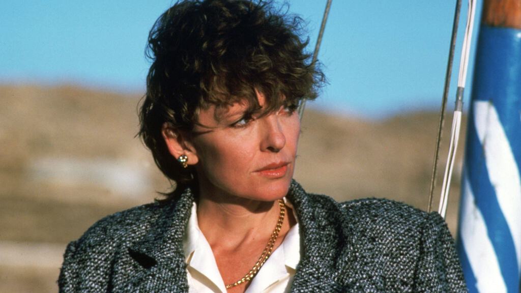 A middle-aged woman with curly brunette hair wearing a tweed blazer and a thoughtful expression, stands outdoors under a clear sky.