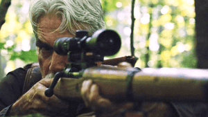 ‘The Man Who Killed Hitler And Then The Bigfoot’ 4K UHD Blu-Ray SteelBook Review – A Rich Role For Sam Elliott An older man aiming a sniper rifle with a scope in a wooded area.