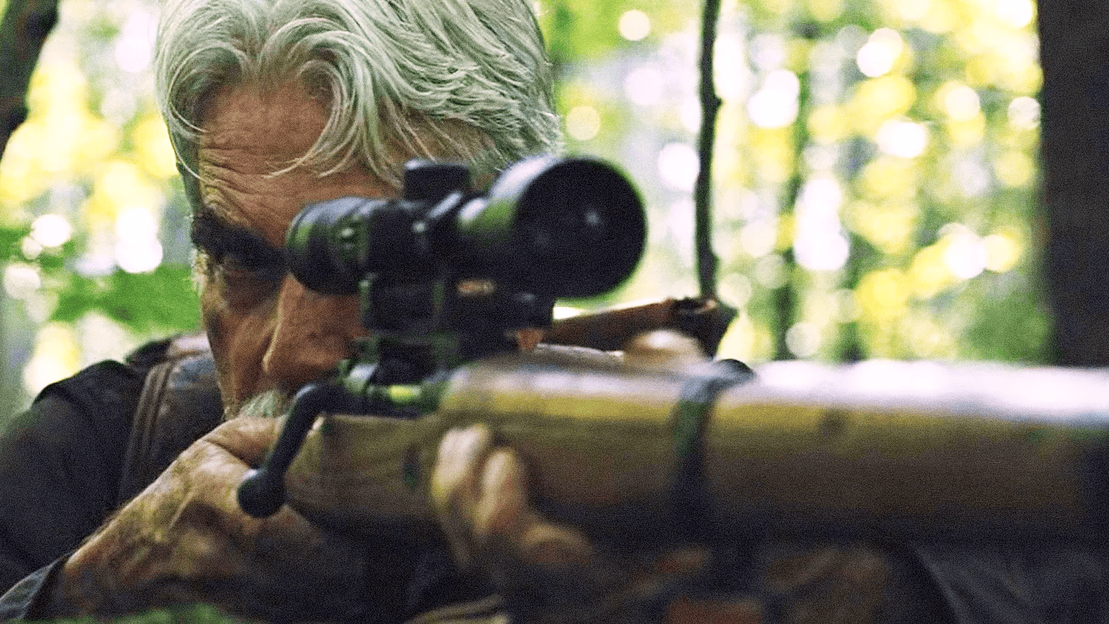 An older man aiming a sniper rifle with a scope in a wooded area.