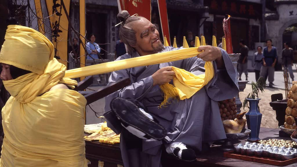 Man in traditional chinese attire playing a stringed instrument at a bustling market, with food vendors and flags in the background.