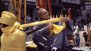 Man in traditional chinese attire playing a stringed instrument at a bustling market, with food vendors and flags in the background.