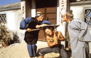 Three actors in a martial arts film scene, dressed in traditional outfits, performing a fight sequence in a village setting.