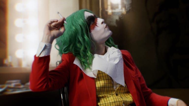 Person in clown makeup with green hair, wearing a red jacket and yellow vest, smoking a cigarette and looking up.