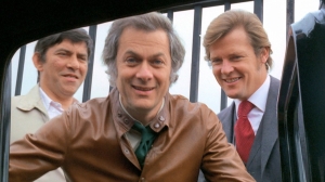 Three men smiling at the camera, one in a brown jacket, another in a red tie, and the third in a dark suit, gathered around an open car window.