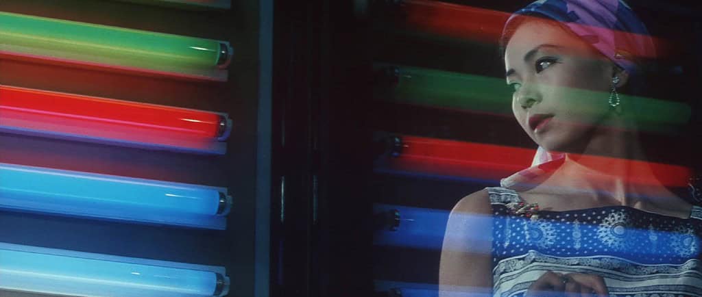 A woman in a colorful headscarf stands contemplatively beside glowing neon lights.