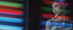 A woman in a colorful headscarf stands contemplatively beside glowing neon lights.