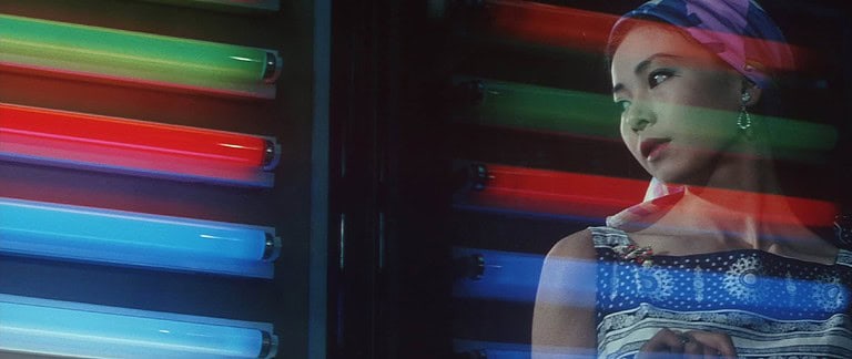 ‘The Shape Of Night’ Blu-Ray Review – A Devastating Downward Spiral A woman in a colorful headscarf stands contemplatively beside glowing neon lights.