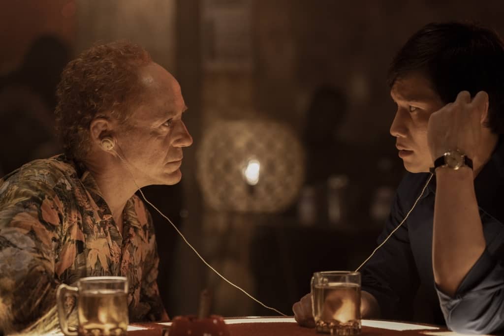 Two men in a tense conversation at a bar, connected by a string tied around their ears, with drinks on the table in a dimly lit setting.