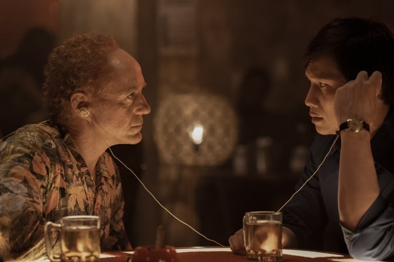 Two men in a tense conversation at a bar, connected by a string tied around their ears, with drinks on the table in a dimly lit setting.
