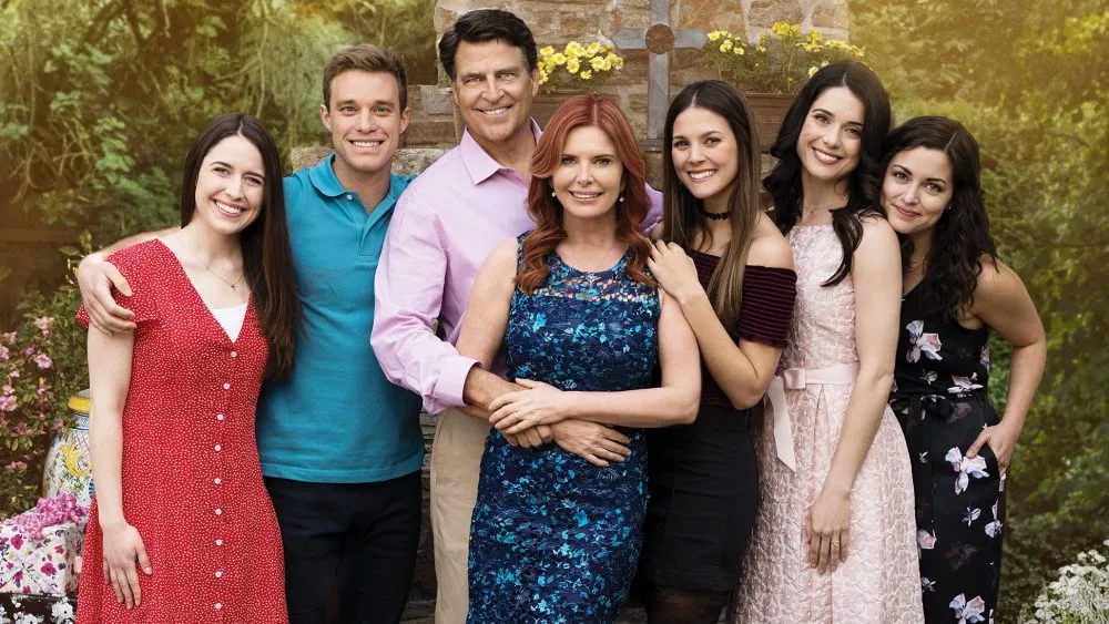 A family of six adults smiling outdoors with a floral background, dressed in casual summer attire.