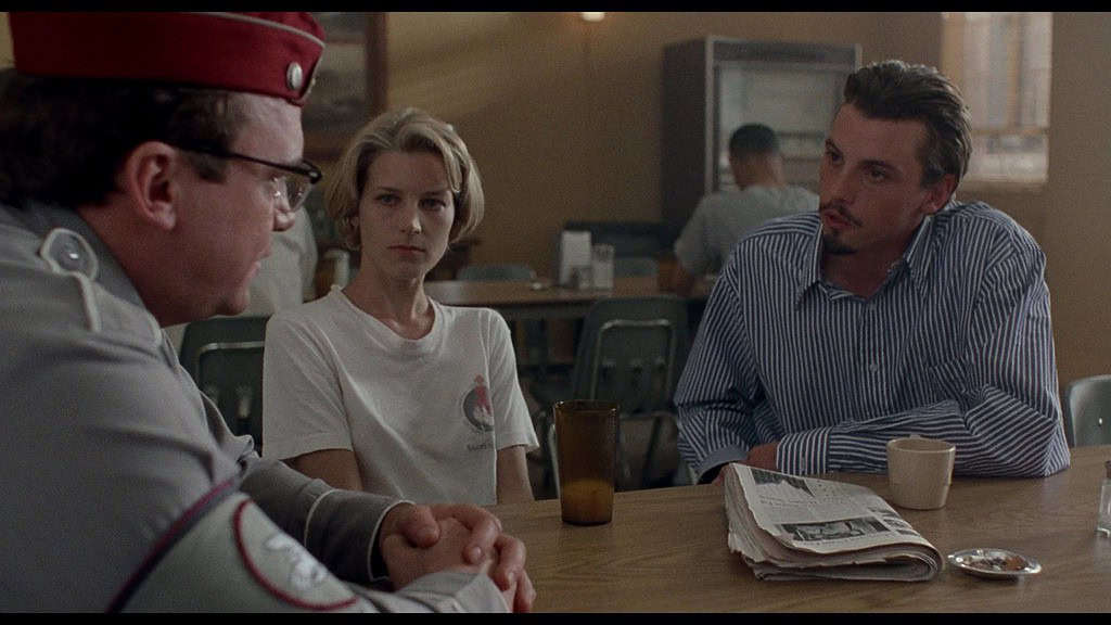 A tense conversation between three people in a diner; a woman and two men, one in a uniform, sitting at a table with a newspaper and drinks.