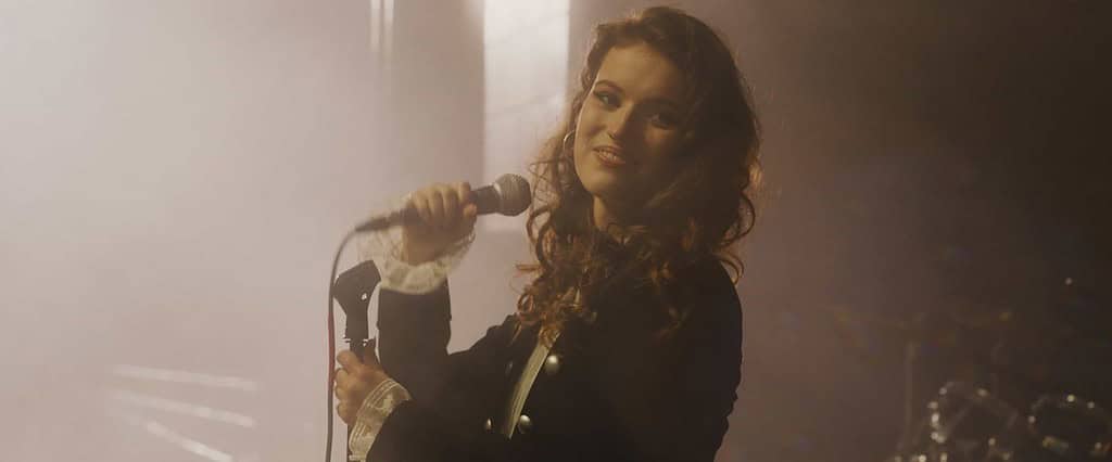 A woman singing on stage with a microphone, highlighted by soft stage lighting with a smoky background.
