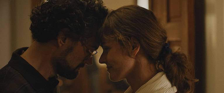‘Unsung Hero’ Review – A Humbling Reminder Of What’s Important A man and woman stand close, facing each other with intense expressions, in a warmly lit indoor setting.