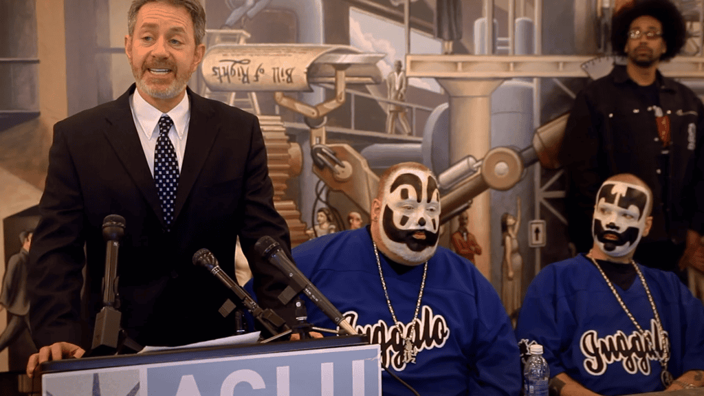 A man in a suit speaks at a podium flanked by two individuals with face paint resembling clowns.