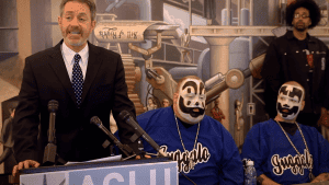 A man in a suit speaks at a podium flanked by two individuals with face paint resembling clowns.