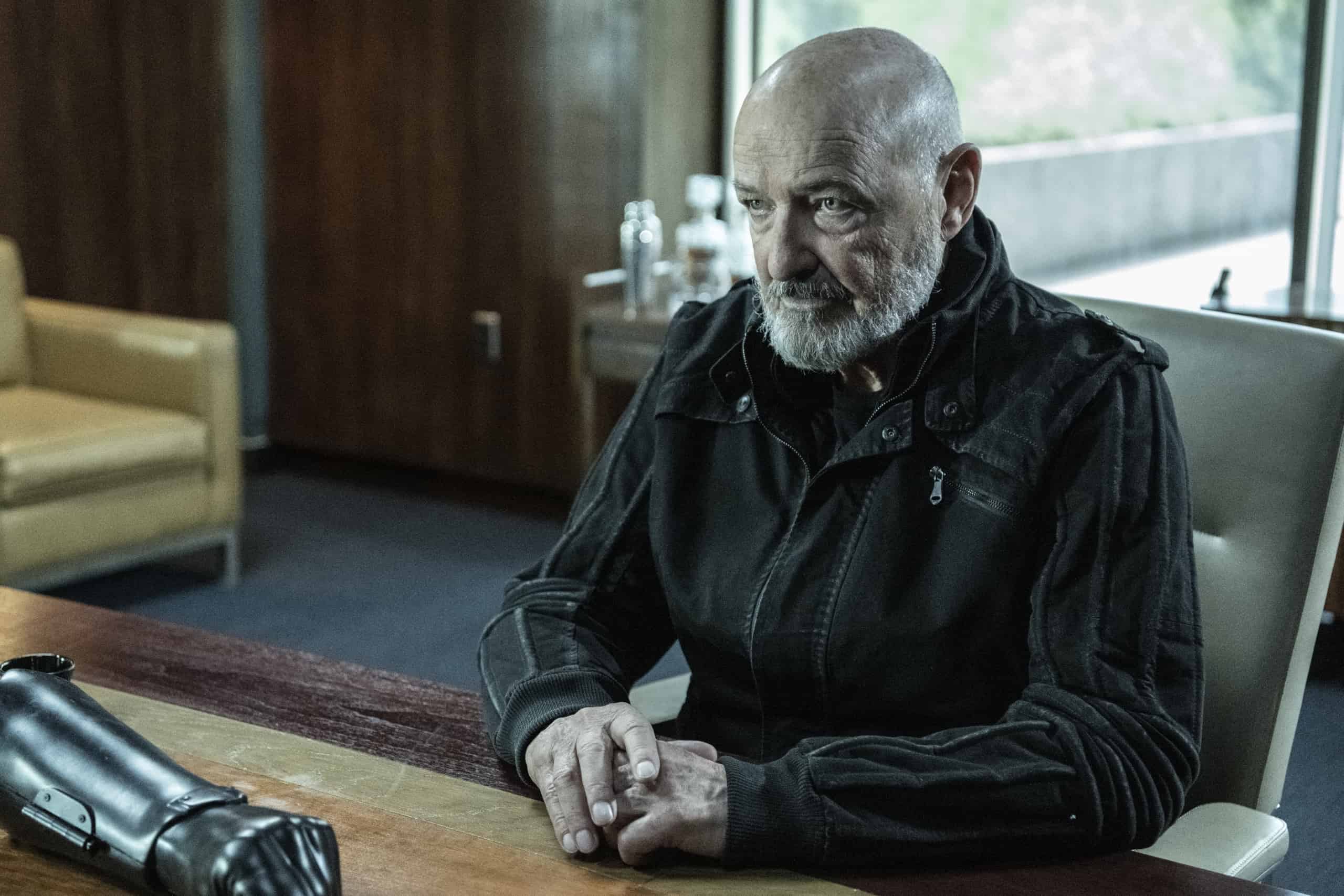 An older man with a beard sitting at a table with a serious expression, with a bionic arm lying in front of him.